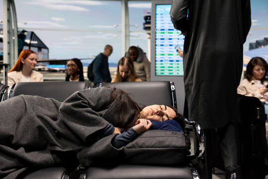 Following a delayed flight, exhausted woman naps with a pillow on airport chairs, resting beside her luggage as she waits through the disruption for another plane to continue the journey.