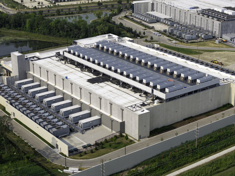 Aerial view of a large data center facility with rows of cooling units on the roof, surrounded by paved roads and green landscape in Illinois, United States.