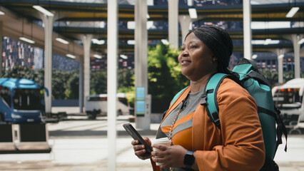 Fototapeta premium Woman enjoys coffee and uses a smartphone in a bus station, carrying a takeaway drink and checking travel information for an intercity coach departure in a close-up scene as a solo passenger.