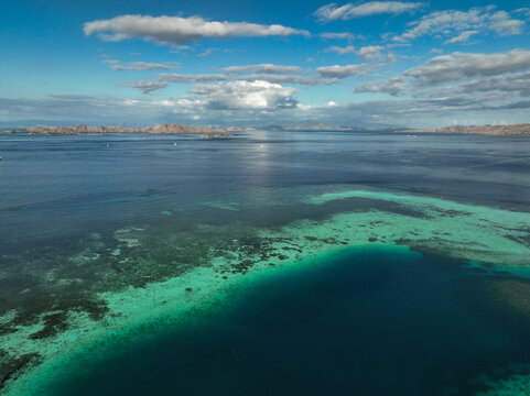 Aerial view of Taka Makasar, a crescent-shaped sandbar surrounded by turquoise coral reefs and deep blue ocean waters under a bright sky in Komodo, Nusa Tenggara Timur, Indonesia.