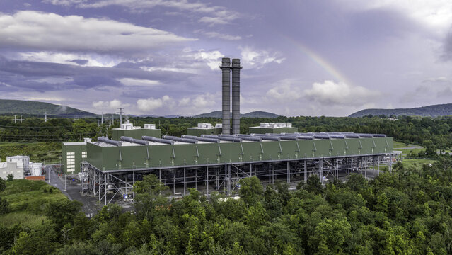 Aerial view of the Cricket Valley Energy Center with exhaust stacks and air-cooled condensers surrounded by forest and a faint rainbow in Dover Plains, New York, United States.
