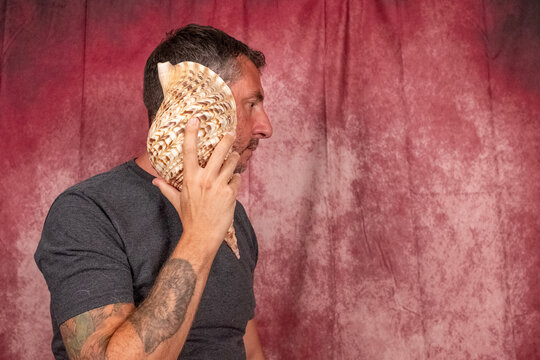 A man holds a large shell to his ear to listen to the sound of the sea. The man is forty-two years old and wears a dark T-shirt