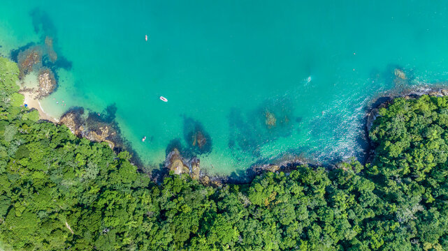 Aerial view of lush coastal forest meeting turquoise waters with small boats floating near the rocky shoreline in Bombinhas, Santa Catarina, Brazil.