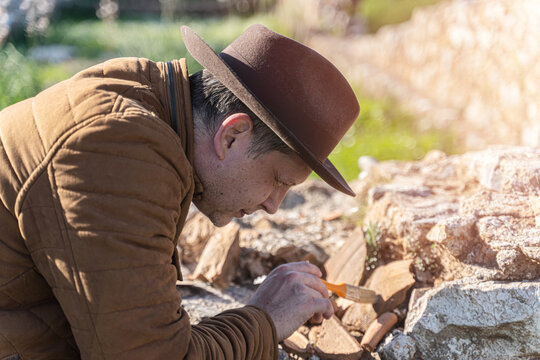 Male archaeologist in hat carefully brushing ancient artifacts at excavation site outdoors