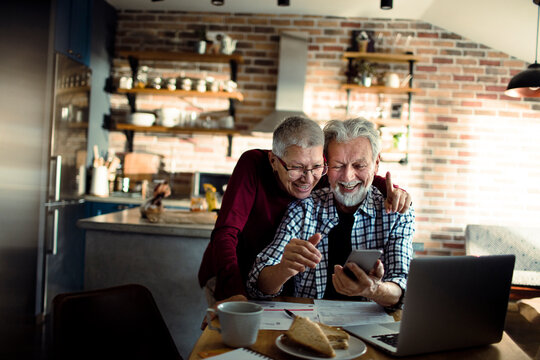 Happy senior couple managing finances on smartphone in home kitchen