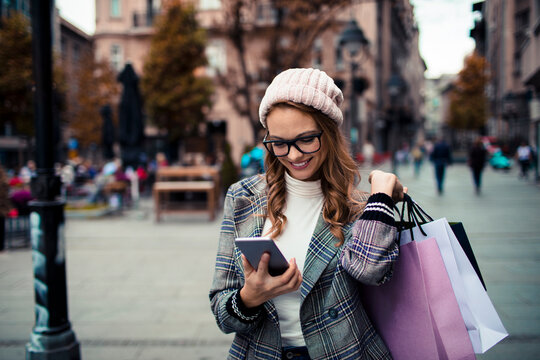 Smiling woman with shopping bags using smartphone on city street