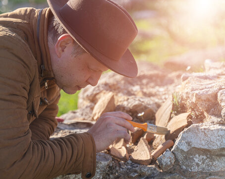 Male archaeologist in hat carefully brushing ancient artifacts at excavation site outdoors