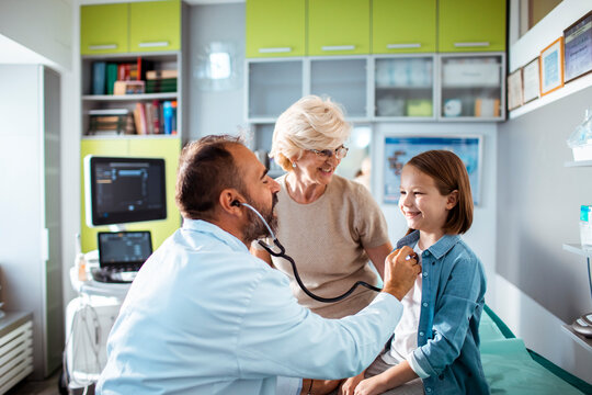 Pediatrician using stethoscope examines smiling girl with grandmother in clinic