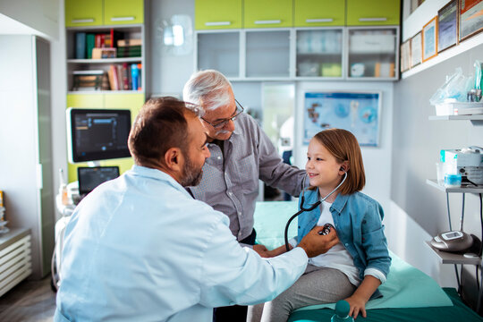 Pediatrician checking girl's heartbeat with grandfather in clinic exam room