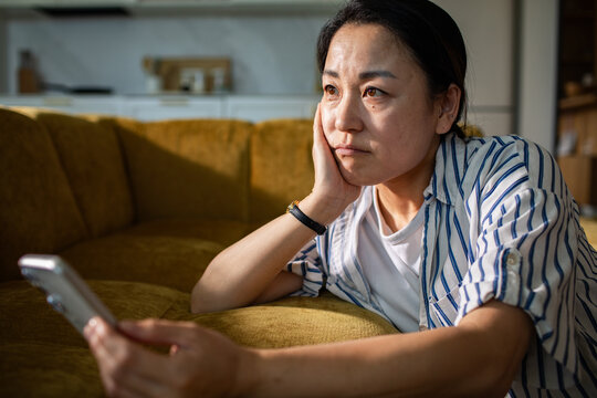 Pensive woman with smartphone on home couch in living room