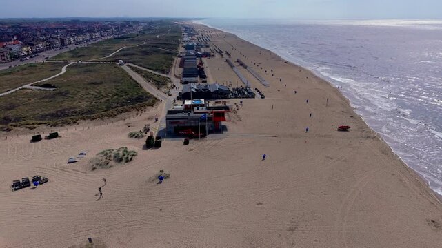 Aerial view video clip of Katwijk aan Zee coastline in Netherlands, featuring lifeguard station, beach pavilions, sand dunes, coastal town, and North Sea horizon on a sunny day