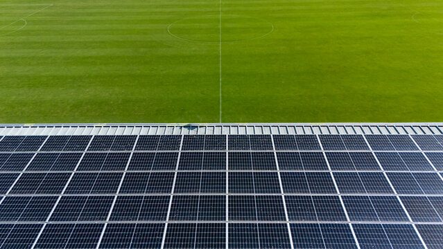 Aerial view of a large solar panel array installed directly adjacent to a green soccer field with white pitch markings in Mill, North Brabant, Netherlands.