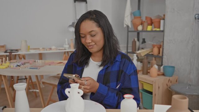 Young woman smiling while scrolling a smartphone with both hands above a clay vase on a pottery table in art studio, wearing a blue plaid shirt; creative focused.