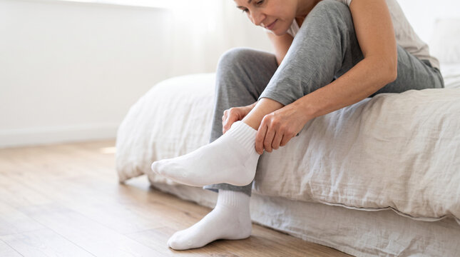 Person putting on socks while sitting on bed with cozy atmosphere and soft light. Individual in casual clothing slips on white socks, enjoying comfort in relaxed home setting.