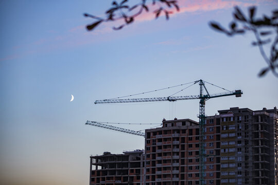Construction site of high-rise apartment building with crane under evening sky and crescent moon
