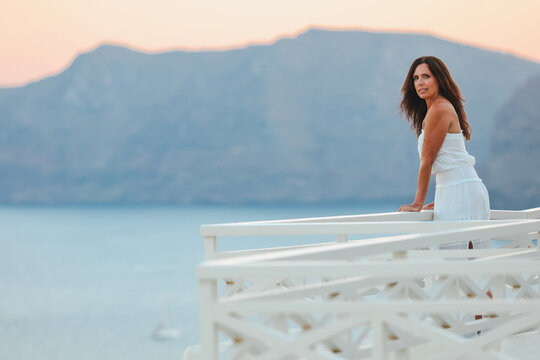 Woman in white dress leaning on a terrace railing overlooking the Aegean sea in Santorini, Greece with soft pastel sky in the background.