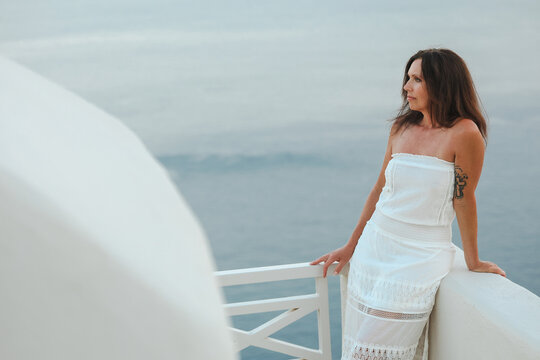 Beautiful, middle aged woman in white dress leaning on a terrace railing overlooking the Aegean sea in Santorini, Greece with a soft pastel sky in the background.