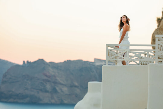 Beautiful, middle aged woman in white dress leaning on a terrace railing overlooking the Aegean sea in Santorini, Greece with a soft pastel sky in the background.