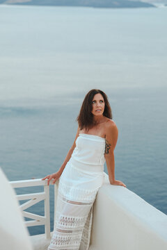 Beautiful, middle aged woman in white dress leaning on a terrace railing overlooking the Aegean sea in Santorini, Greece with a soft pastel sky in the background.