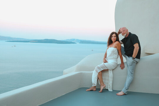 Middle‑aged couple enjoying a quiet moment together on a white terrace overlooking the Aegean sea at Santorini, Greece during sunset.