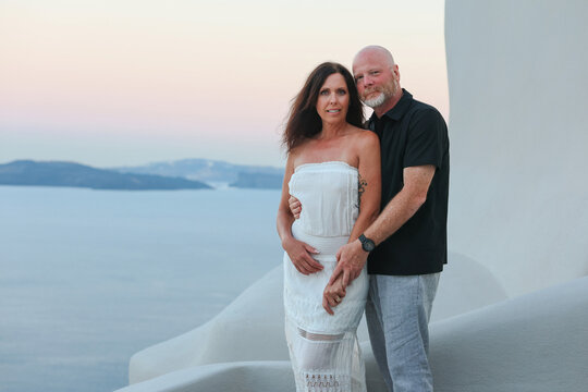 Middle‑aged couple enjoying a quiet moment together on a white terrace overlooking the Aegean sea at Santorini, Greece during sunset.