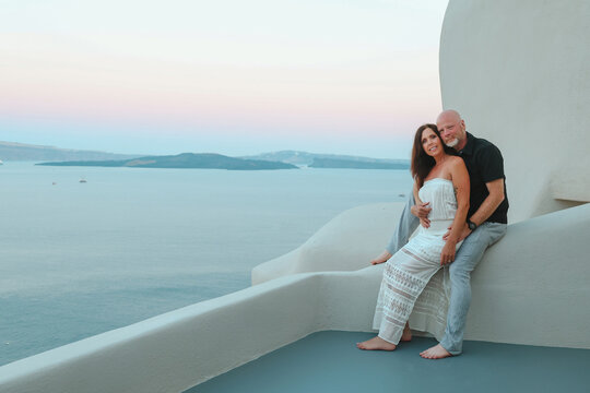 Middle‑aged couple enjoying a quiet moment together on a white terrace overlooking the Aegean sea at Santorini, Greece during sunset.