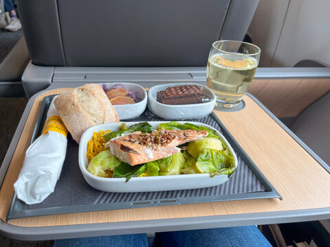 Premium train business class meal tray with grilled salmon, greens, bread, dessert and white wine glass on wooden fold-down table