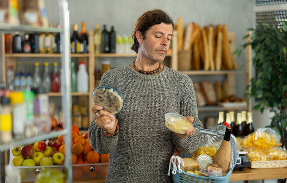 Focused man with shopping basket trying to make choice holding two sorts of herring in grocery store with varied stock