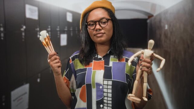Young woman holds paintbrushes and wooden mannequin in studio corridor, wearing yellow beret and glasses; creative confidence.