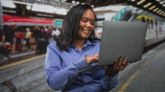 Woman typing on a laptop in a busy station building, smiling and holding the screen while commuters pass on platforms; happy productivity.