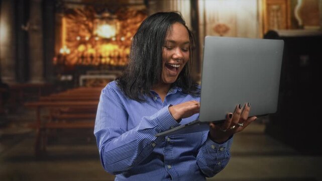 African american woman laughing and typing on laptop while holding it with one hand, hand to mouth surprise gesture, in an ornate church interior with wooden pews and altar; delight connection.
