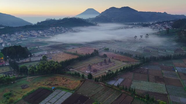 Aerial view of Candi Arjuna temple complex surrounded by mist and agricultural fields at sunrise in Dieng, Indonesia.