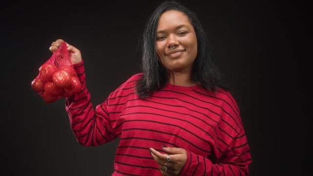 Young african american woman wearing red striped shirt holds a net bag of tomatoes up with one hand and smiles toward camera in studio; simple contentment.