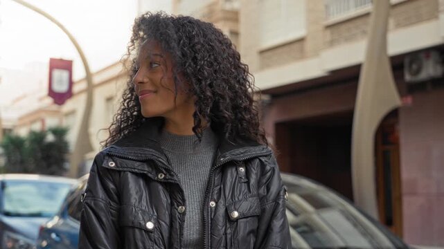 Teenage woman with curly hair smiles and looks left wearing a black puffer jacket on a city street near parked cars; carefree joy.