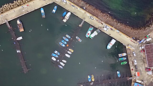 Aerial view of a small working fishing boat moored at a harbor pier, surrounded by seagulls, fishing equipment, parked vehicles and rows of colorful boats along the waterfront.