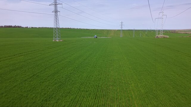 Aerial flight following a blue agricultural sprayer with long extended booms across a green field, with electricity pylons and parallel tracks ahead.