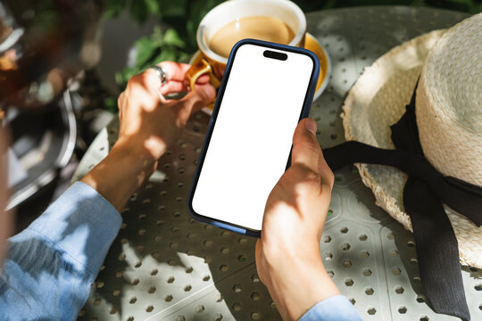 Personal perspective view of a woman sitting at an outdoor cafe holding a mobile phone with a blank screen and drinking a cup of white coffee on a summer day