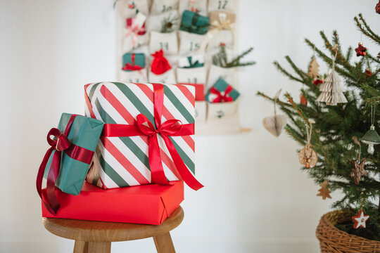 Traditional Christmas advent calendar with assorted wrapped gifts hanging on a white wall next to a Christmas tree and wooden stool with a three larger wrapped Christmas gifts