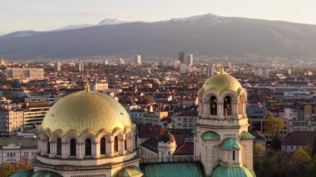 Aerial golden hour view of Alexander Nevsky Cathedral in Sofia, Bulgaria, highlighting its monumental golden dome, Orthodox cross, green-roofed towers and surrounding city center architecture.