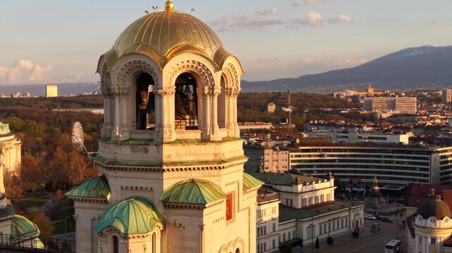 Aerial golden hour view of Alexander Nevsky Cathedral in Sofia, Bulgaria, highlighting its monumental golden dome, Orthodox cross, green-roofed towers and surrounding city center architecture.