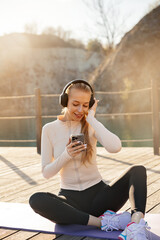 Fototapeta premium Pretty woman enjoying music after outdoor workout. Portrait of female model sitting on yoga mat with smartphone. Wellness concept.