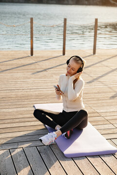 Beautiful woman listening to music after yoga workout. Beauty woman relaxing on fitness mat with headphones outdoors. Wellness concept.