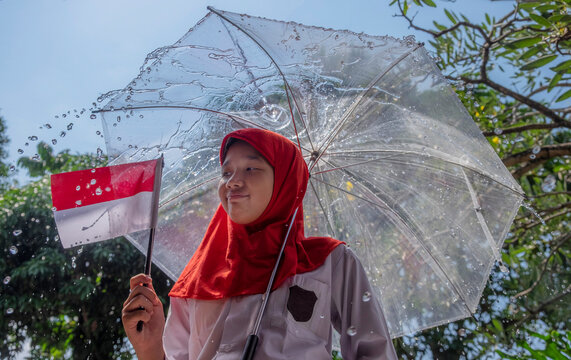 Portrait of a smiling Teenage Girl in a Hijab standing outdoors holding an Indonesian flag and an umbrella