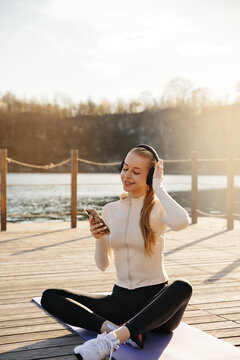 Beauty woman enjoying music after outdoor workout. Attractive woman relaxing on yoga mat with wireless headphones. Wellness concept.