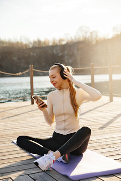 Beautiful woman relaxing after yoga while listening to music. Young woman sitting on fitness mat using smartphone outdoors. Active lifestyle concept.