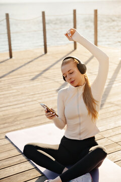 Pretty woman raising arm while listening to favorite music. Portrait of female model holding smartphone after yoga. Fitness concept.