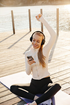 Young woman happily checking smartphone after yoga session. Attractive woman wearing headphones in warm sunlight outdoors. Wellness concept.
