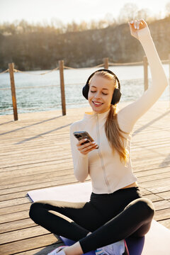 Beautiful woman celebrating workout while holding smartphone outdoors. Young woman smiling with headphones on yoga mat. Fitness lifestyle concept.
