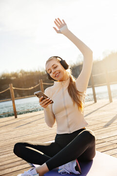 Beauty woman relaxing with headphones after outdoor workout. Attractive woman checking smartphone in warm golden sunlight. Wellness concept.