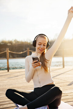 Beautiful woman smiling while using smartphone on yoga mat. Young woman enjoying music through headphones outdoors. Fitness lifestyle concept.